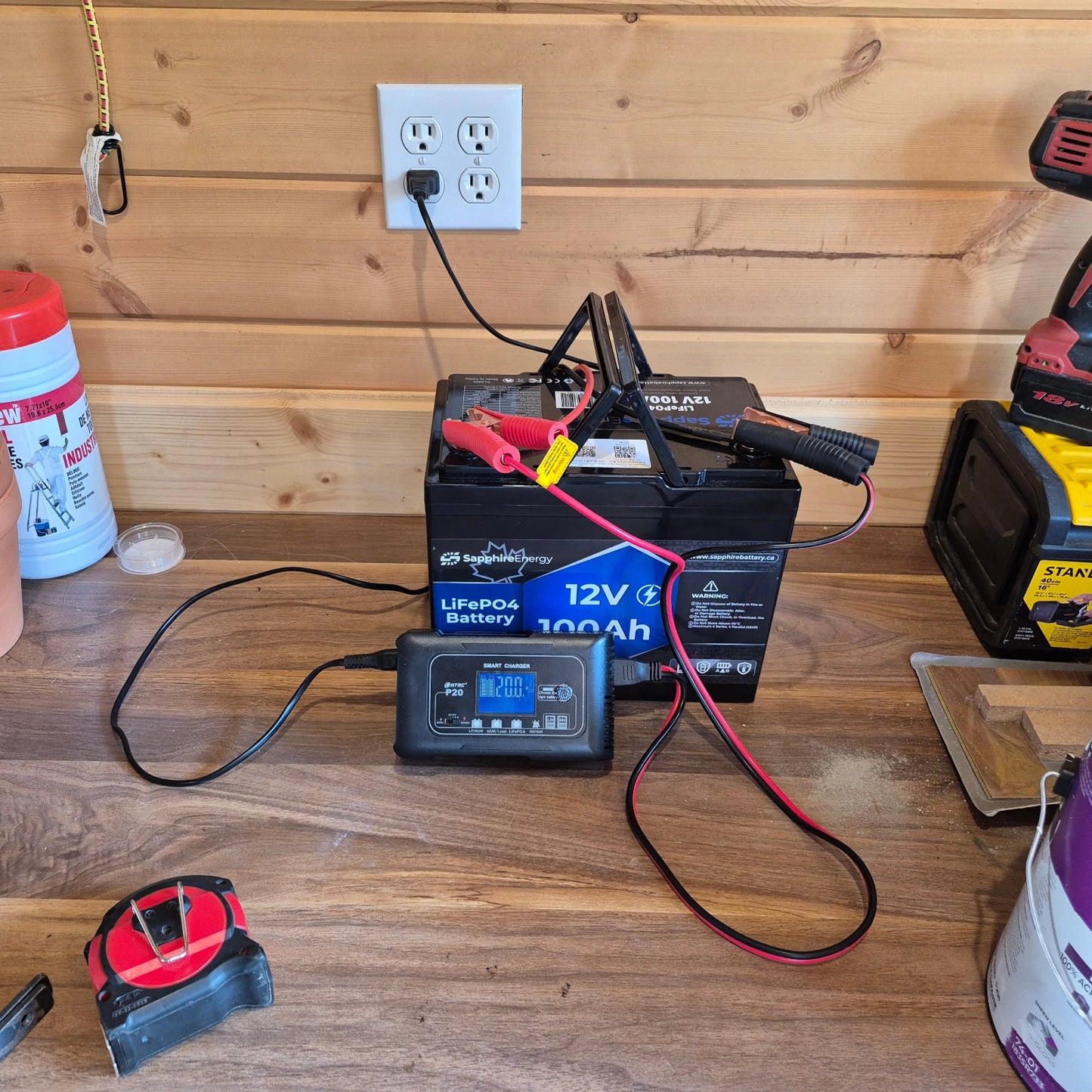 Battery charger connected to a battery on a wooden surface with a wall outlet in the background.